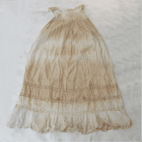 Light cream child’s dress laid flat, featuring gathered fabric and decorative horizontal bands near the hem, with extensive brown staining throughout.